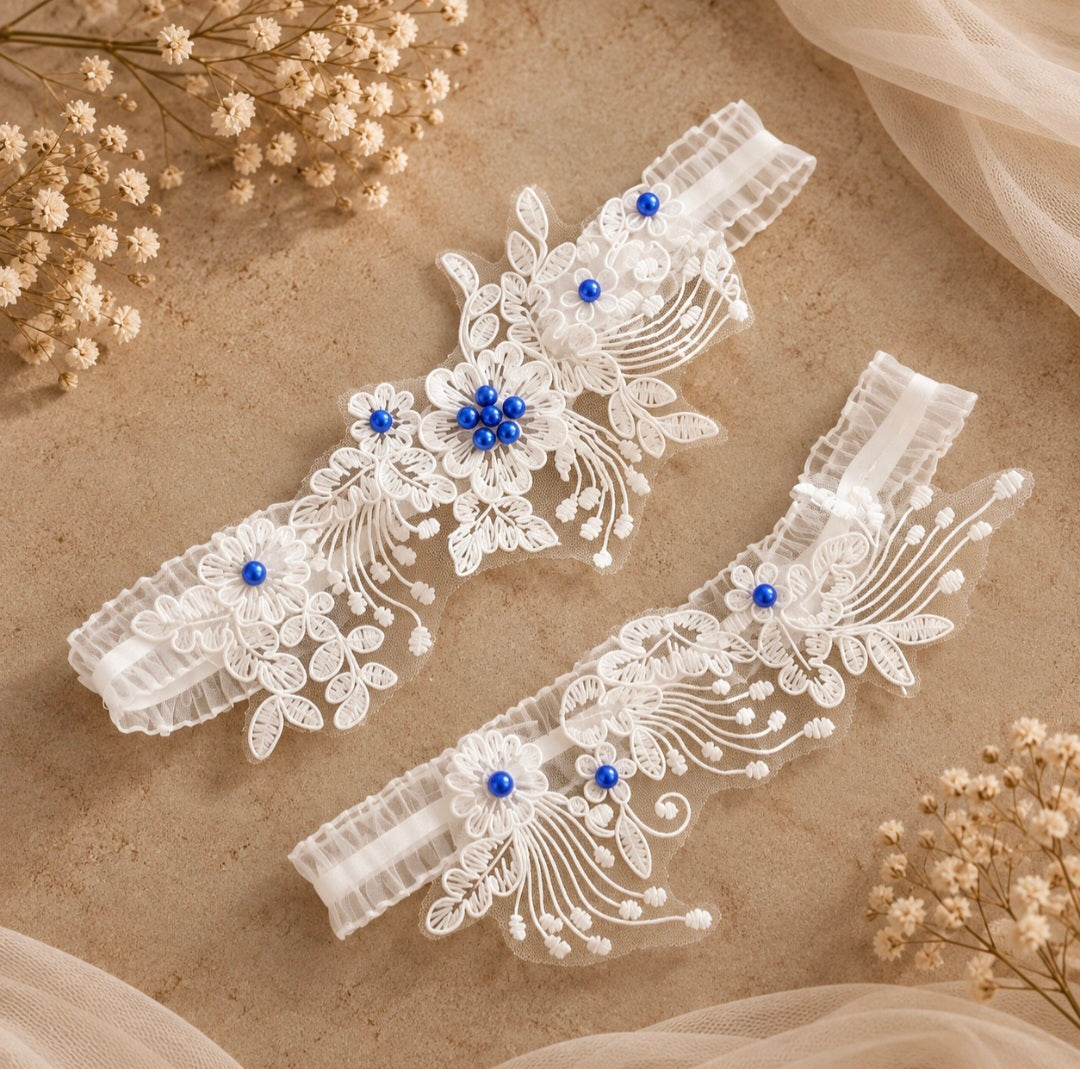 Two white lace bridal garter belts with blue floral bead accents and pearl details, arranged on a beige textured surface with dried flowers and sheer fabric.