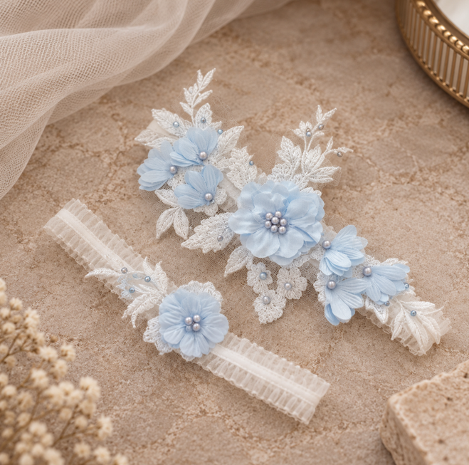 Two white lace garter belts decorated with light blue fabric flowers, white lace leaves, and small pearl-like beads, arranged on a textured beige surface with soft tulle and dried flowers.