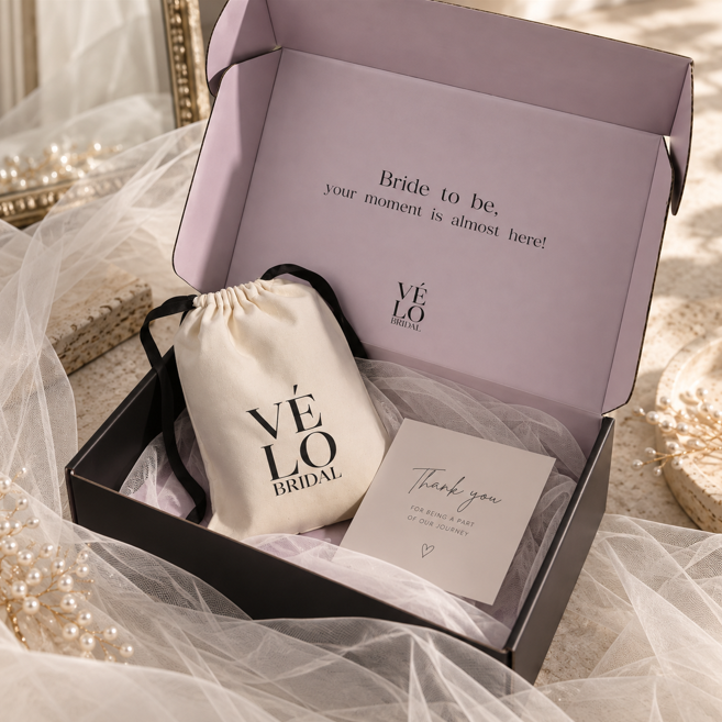 A bridal gift box set from Vélo Bridal, featuring a lavender interior insert with the message 'Bride to be, your moment is almost here!', a cream drawstring pouch with black branding, and a thank you card nestled in tulle inside a black box.
