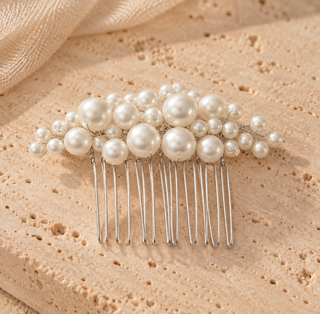 A pearl hair comb featuring large and small white faux pearls on a silver metal comb base, displayed on a textured beige stone surface.