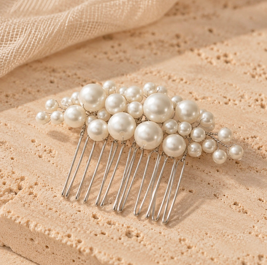 A pearl hair comb accessory with large and small white faux pearls on silver-tone metal prongs, displayed on a textured beige stone surface with soft cream fabric in the background.