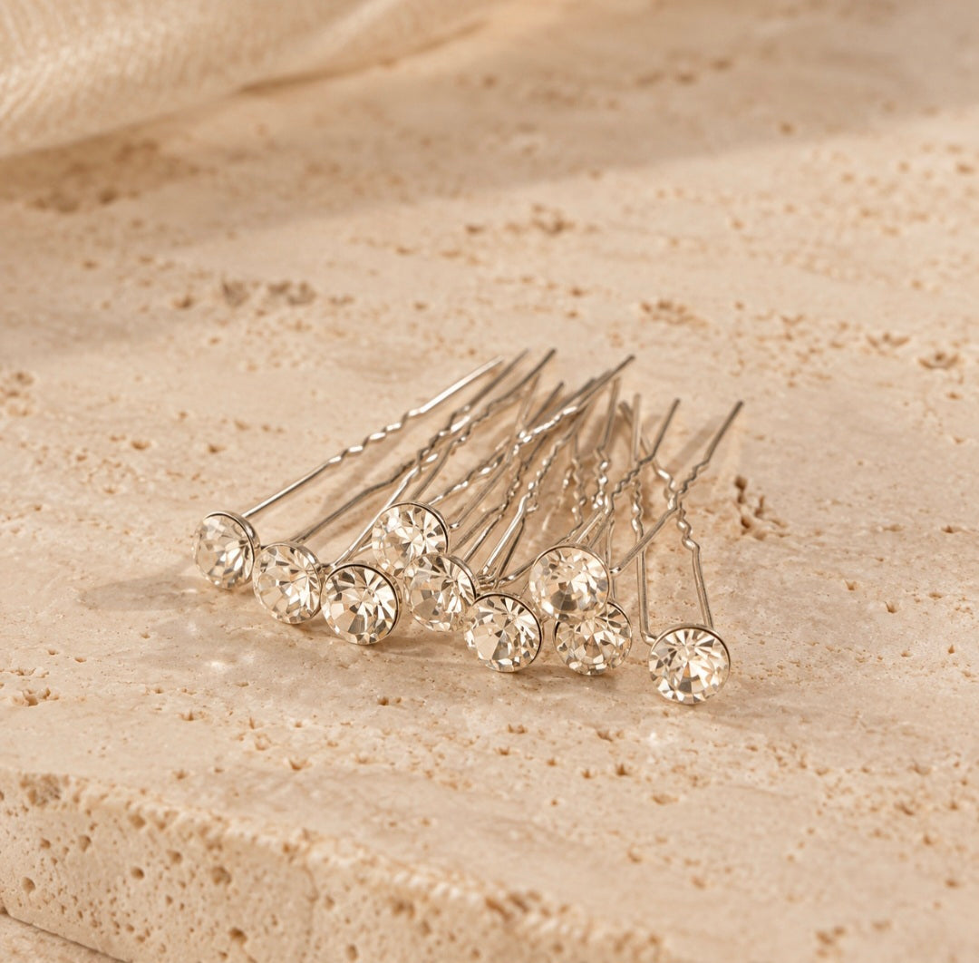 A set of silver hair pins with clear faceted crystal ball tops, arranged on a textured beige stone surface.