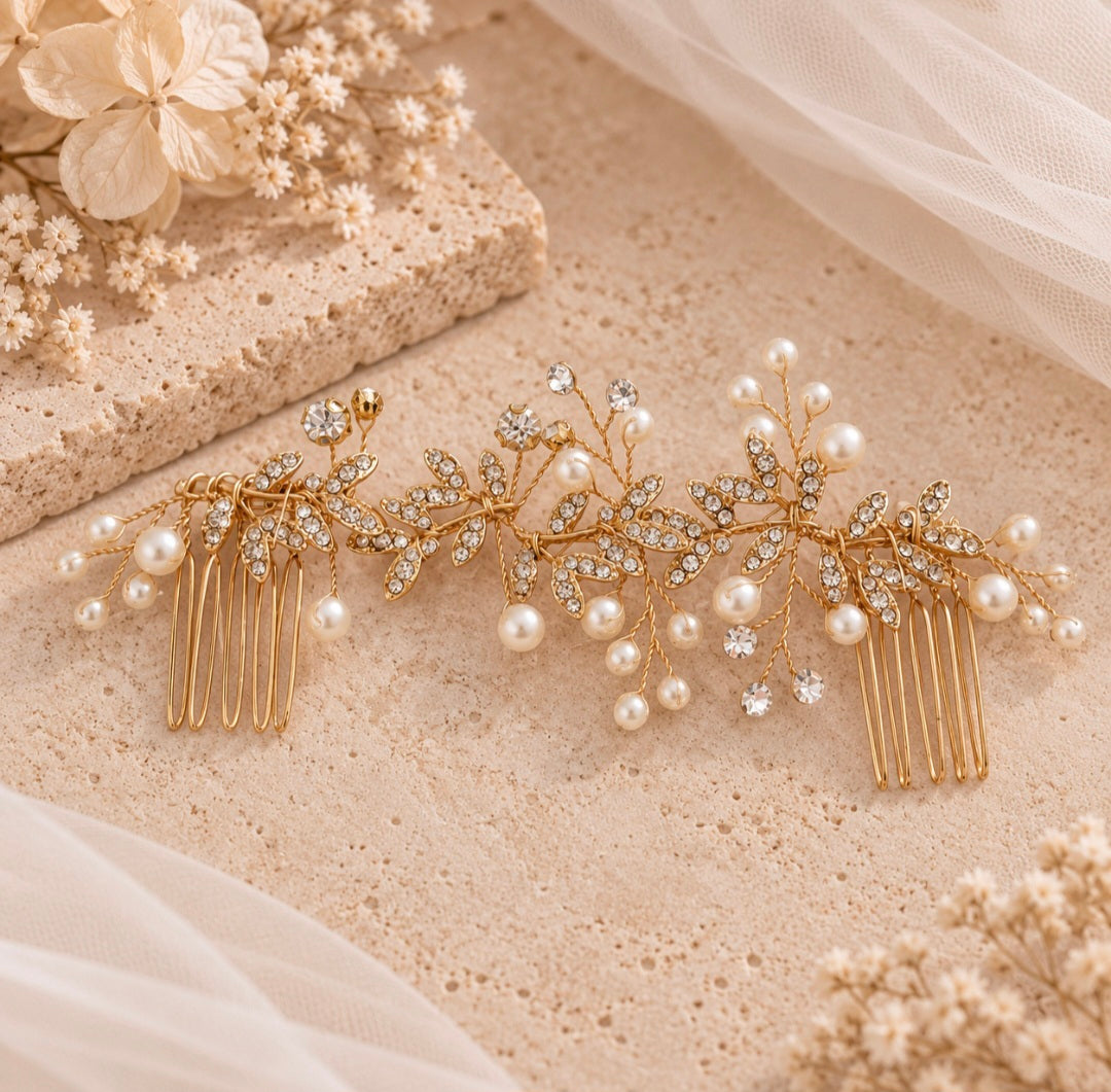 A gold leaf-shaped hair comb accessory decorated with faux pearls and clear rhinestones, displayed on a textured beige surface with soft tulle and dried floral props.