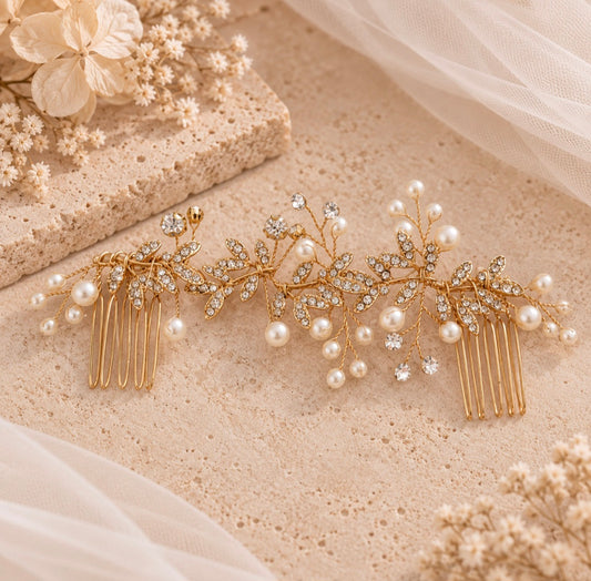 A gold leaf-shaped hair comb accessory decorated with faux pearls and clear rhinestones, displayed on a textured beige surface with soft tulle and dried floral props.