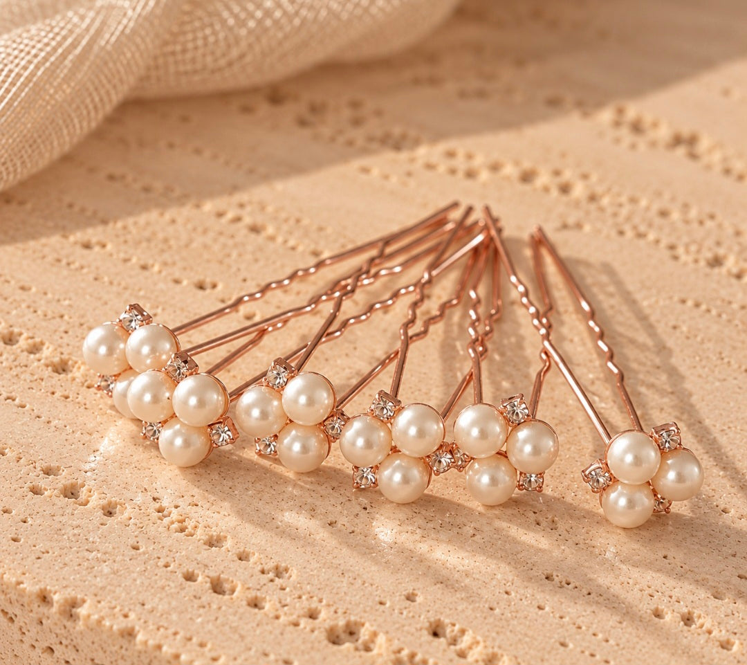 Six rose gold hair pins with clusters of white faux pearls and small clear rhinestone accents, arranged on a textured beige stone surface.