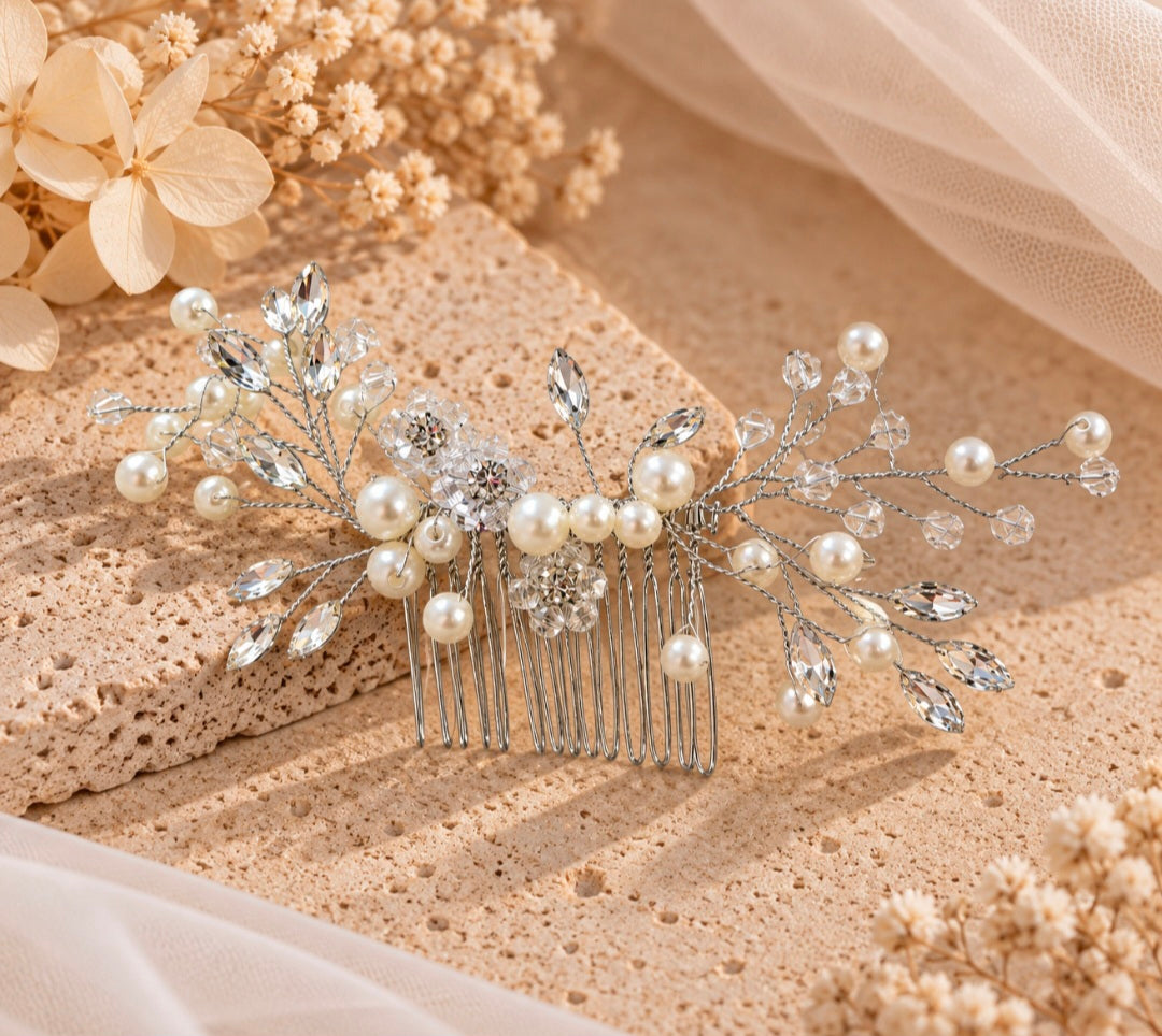 A silver bridal hair comb decorated with faux pearls, clear rhinestones, and crystal branches, styled on a textured beige stone slab with dried flowers and sheer fabric.