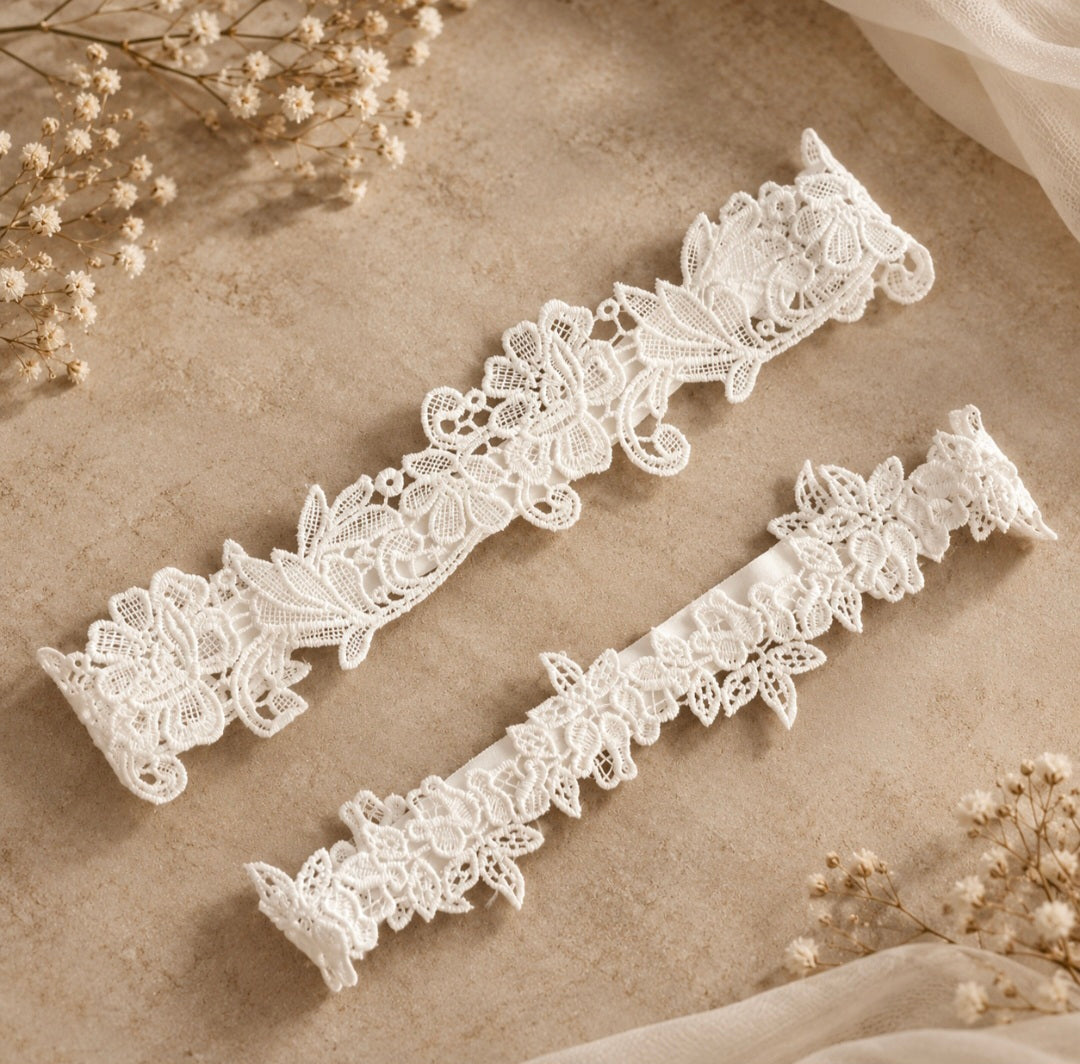 Two white lace garter belts with floral and leaf patterns, displayed diagonally on a textured beige surface with dried baby's breath flowers.