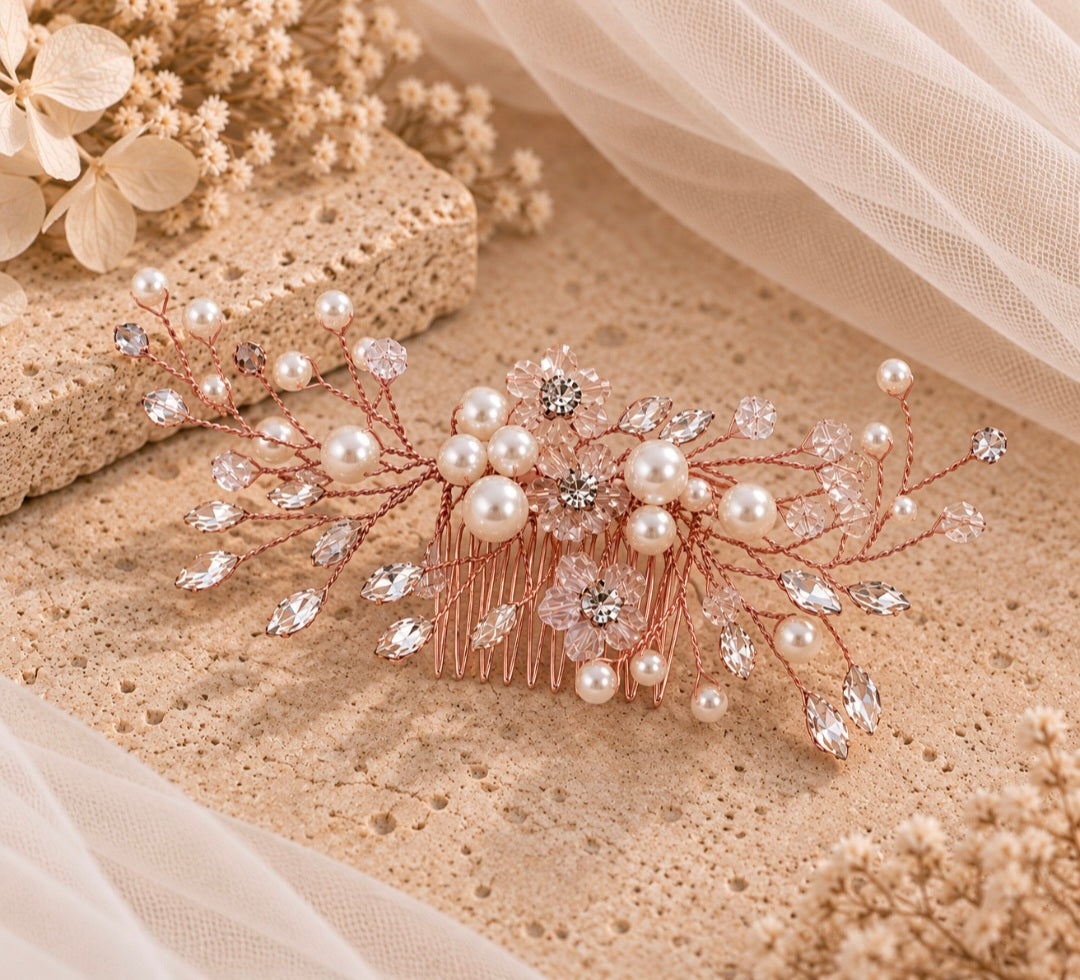 A bridal hair comb featuring rose gold wire branches adorned with faux pearls, clear rhinestones, and crystal drops, styled on a textured beige stone surface with soft white tulle fabric.