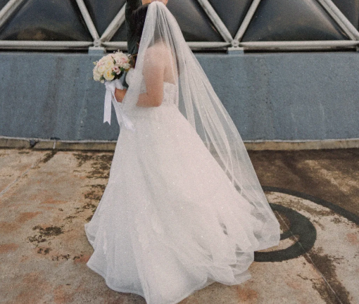 A bride in a sparkling white strapless wedding gown with a long sheer veil, holding a pastel bouquet and photographed from behind against a gray wall and glass roof structure.